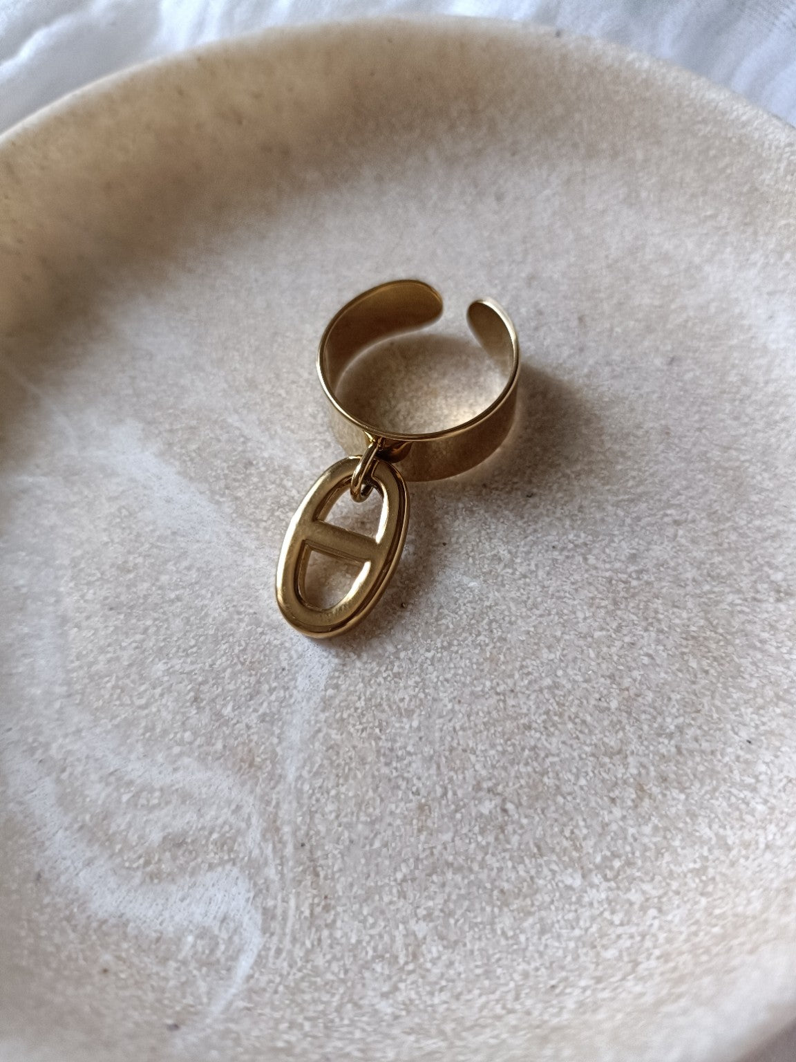 Bague dorée en acier inoxydable avec breloque maille marine de 2 cm posée sur une coupelle beige
